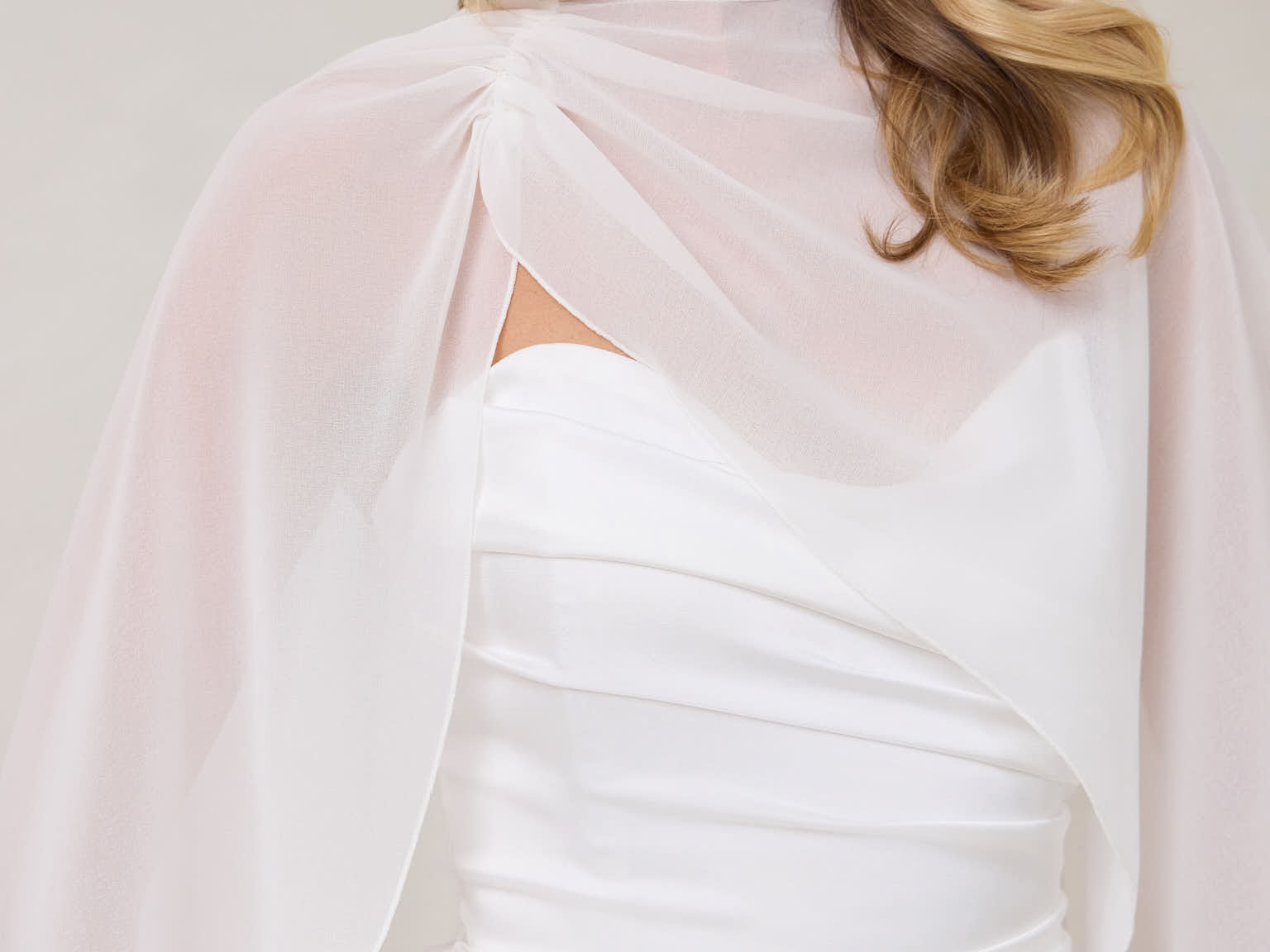 A woman is standing against a white backdrop. She's wearing a white bridal dress with a chiffon cape over her shoulders. The cape is gathered slightly on the left side and is nearly fingertip length. This is the Cleo Cape.
