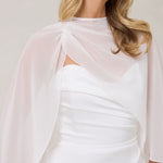 A woman is standing against a white backdrop. She's wearing a white bridal dress with a chiffon cape over her shoulders. The cape is gathered slightly on the left side and is nearly fingertip length. This is the Cleo Cape.