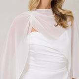 A woman is standing against a white backdrop. She's wearing a white bridal dress with a chiffon cape over her shoulders. The cape is gathered slightly on the left side and is nearly fingertip length. This is the Cleo Cape.