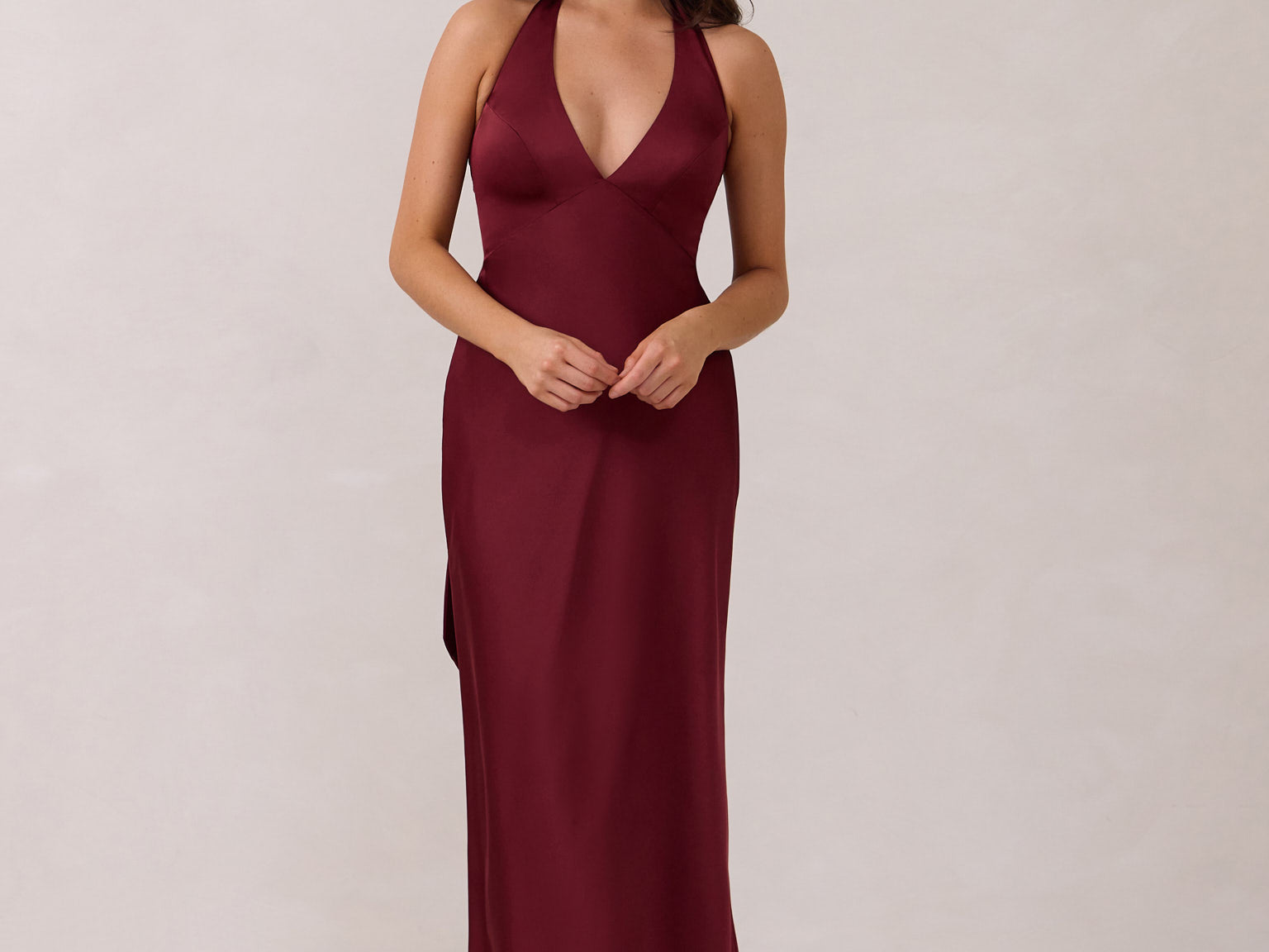 A woman is standing against a white backdrop. She's wearing a burgundy, satin, tea-length bridesmaid dress. The dress has a halter neckline with a scarf detail. The dress is fitted through the bodice and falls nicely over the hips. Perfect to wear as a bridesmaid dress or as a wedding guest dress or any special event. 
