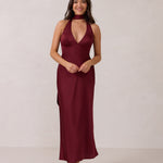 A woman is standing against a white backdrop. She's wearing a burgundy, satin, tea-length bridesmaid dress. The dress has a halter neckline with a scarf detail. The dress is fitted through the bodice and falls nicely over the hips. Perfect to wear as a bridesmaid dress or as a wedding guest dress or any special event. 