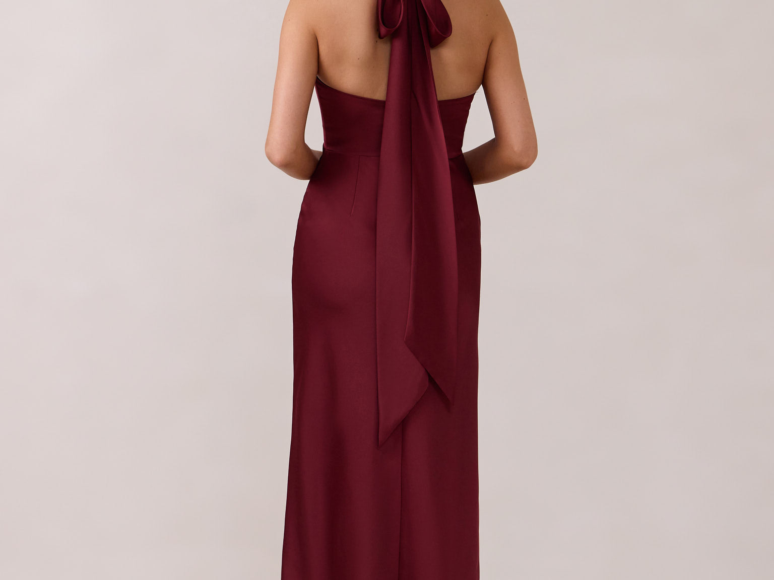 A woman is standing against a white backdrop facing the back. She's wearing a burgundy, satin, tea-length bridesmaid dress. The dress has a halter neckline with a scarf detail. The dress is fitted through the bodice and falls nicely over the hips. Perfect to wear as a bridesmaid dress or as a wedding guest dress or any special event. 