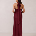 A woman is standing against a white backdrop facing the back. She's wearing a burgundy, satin, tea-length bridesmaid dress. The dress has a halter neckline with a scarf detail. The dress is fitted through the bodice and falls nicely over the hips. Perfect to wear as a bridesmaid dress or as a wedding guest dress or any special event. 
