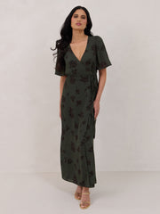 A woman is standing against a white backdrop wearing a dark green wrap dress with a subtle black floral print. The dress has a deep V neckline and loose sleeves. This is the Sherwood Dress in Esme.
