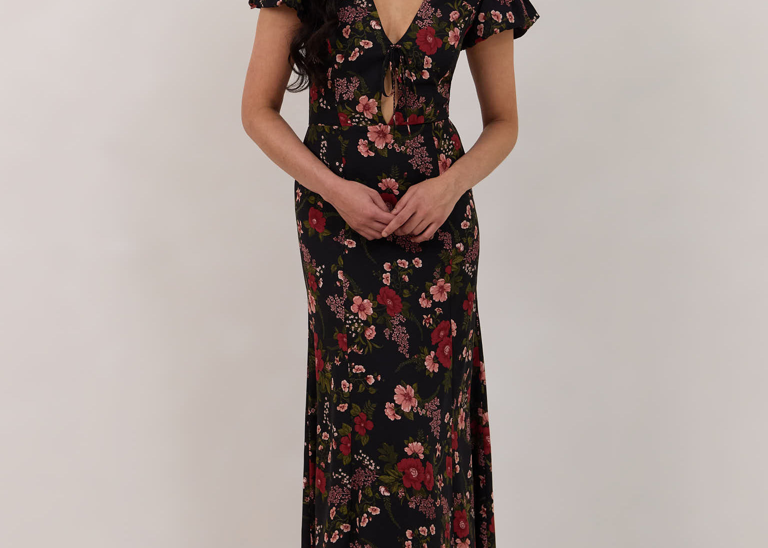 A woman is wearing the Beatrice Dress in Zinnia. This is a black dress with a beautiful red and pink floral print. The dress has a slightly puffed sleeve, and a full back with a keyhole detail. The front has a deep-V cut with a delicate tie at the bust and another keyhole detail down to the waistline. This is a bra-friendly dress that is ankle length. 