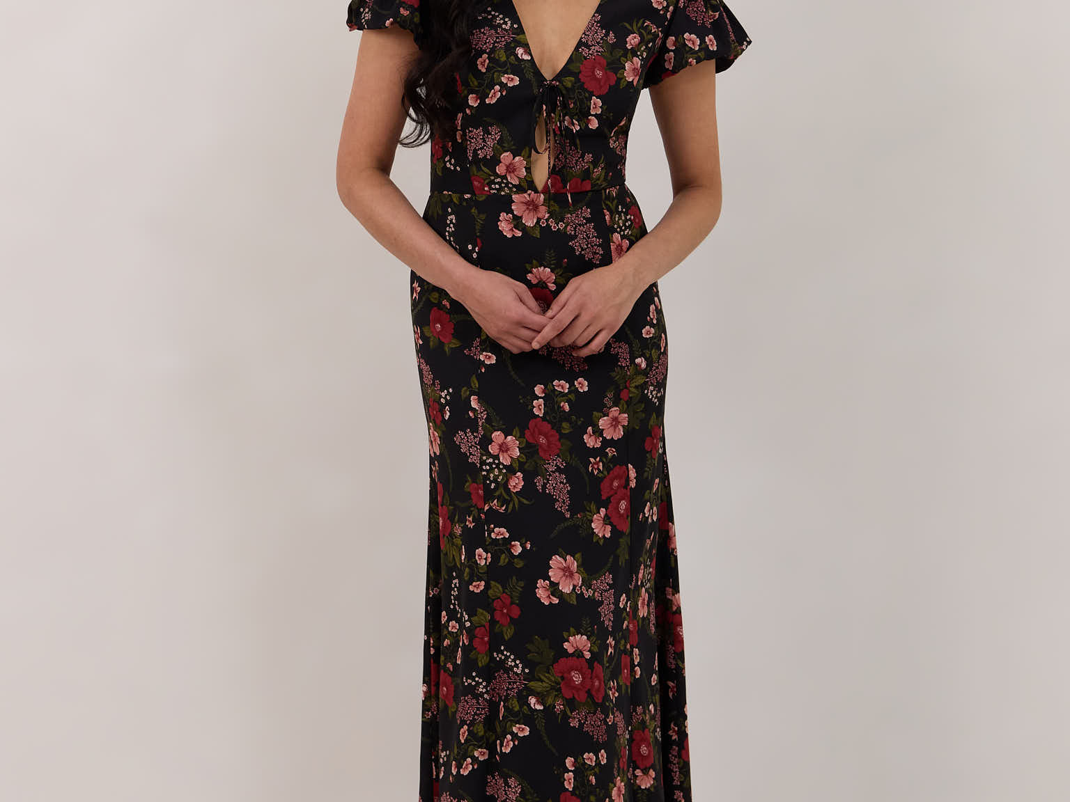A woman is wearing the Beatrice Dress in Zinnia. This is a black dress with a beautiful red and pink floral print. The dress has a slightly puffed sleeve, and a full back with a keyhole detail. The front has a deep-V cut with a delicate tie at the bust and another keyhole detail down to the waistline. This is a bra-friendly dress that is ankle length. 