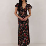 A woman is wearing the Beatrice Dress in Zinnia. This is a black dress with a beautiful red and pink floral print. The dress has a slightly puffed sleeve, and a full back with a keyhole detail. The front has a deep-V cut with a delicate tie at the bust and another keyhole detail down to the waistline. This is a bra-friendly dress that is ankle length. 