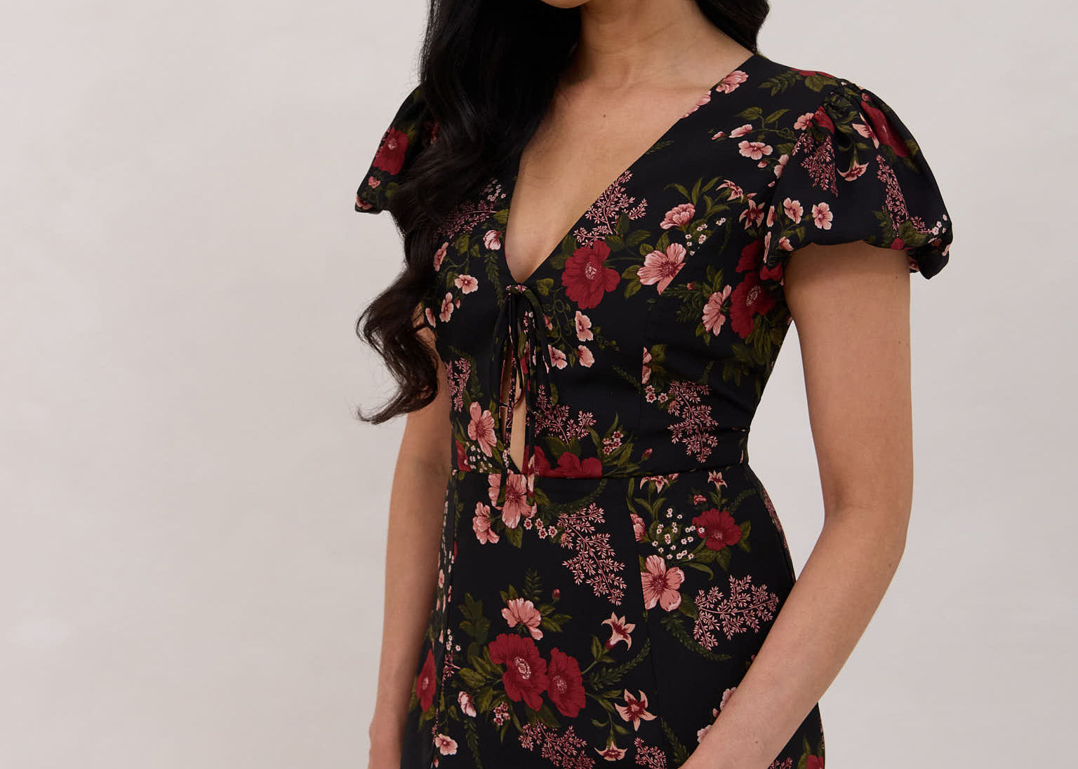 A woman is wearing the Beatrice Dress in Zinnia. This is a black dress with a beautiful red and pink floral print. The dress has a slightly puffed sleeve, and a full back with a keyhole detail. The front has a deep-V cut with a delicate tie at the bust and another keyhole detail down to the waistline. This is a bra-friendly dress that is ankle length. 