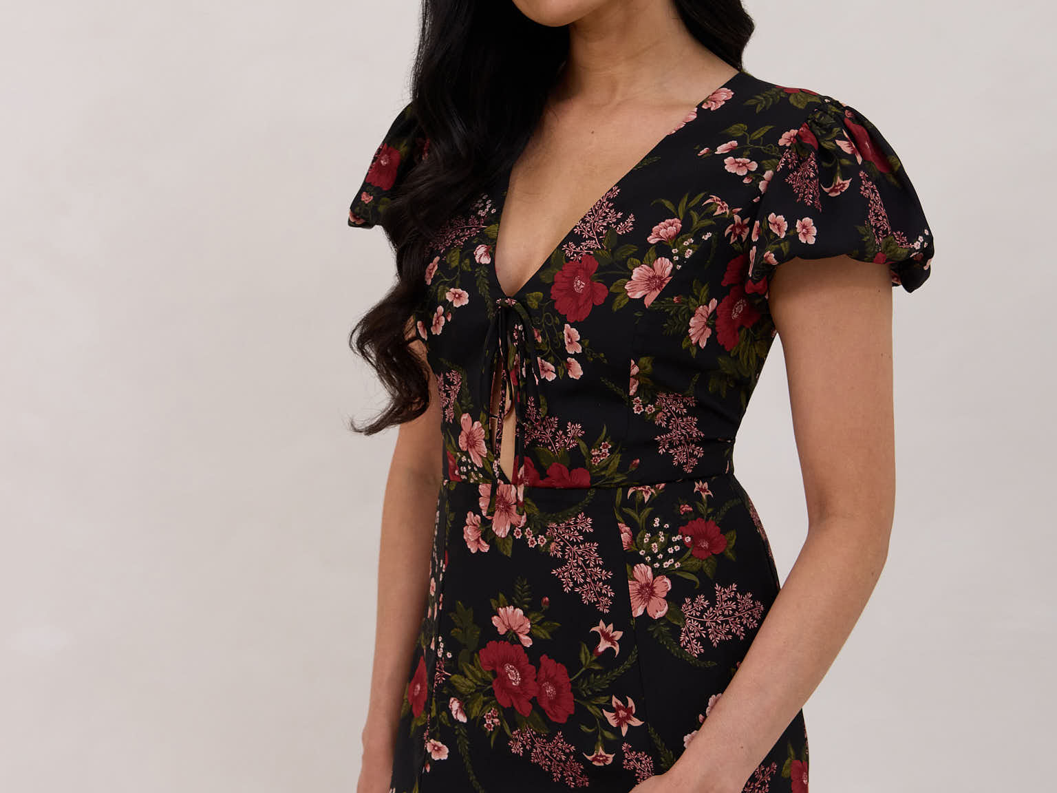 A woman is wearing the Beatrice Dress in Zinnia. This is a black dress with a beautiful red and pink floral print. The dress has a slightly puffed sleeve, and a full back with a keyhole detail. The front has a deep-V cut with a delicate tie at the bust and another keyhole detail down to the waistline. This is a bra-friendly dress that is ankle length. 