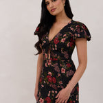 A woman is wearing the Beatrice Dress in Zinnia. This is a black dress with a beautiful red and pink floral print. The dress has a slightly puffed sleeve, and a full back with a keyhole detail. The front has a deep-V cut with a delicate tie at the bust and another keyhole detail down to the waistline. This is a bra-friendly dress that is ankle length. 