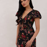 A woman is wearing the Beatrice Dress in Zinnia. This is a black dress with a beautiful red and pink floral print. The dress has a slightly puffed sleeve, and a full back with a keyhole detail. The front has a deep-V cut with a delicate tie at the bust and another keyhole detail down to the waistline. This is a bra-friendly dress that is ankle length. 