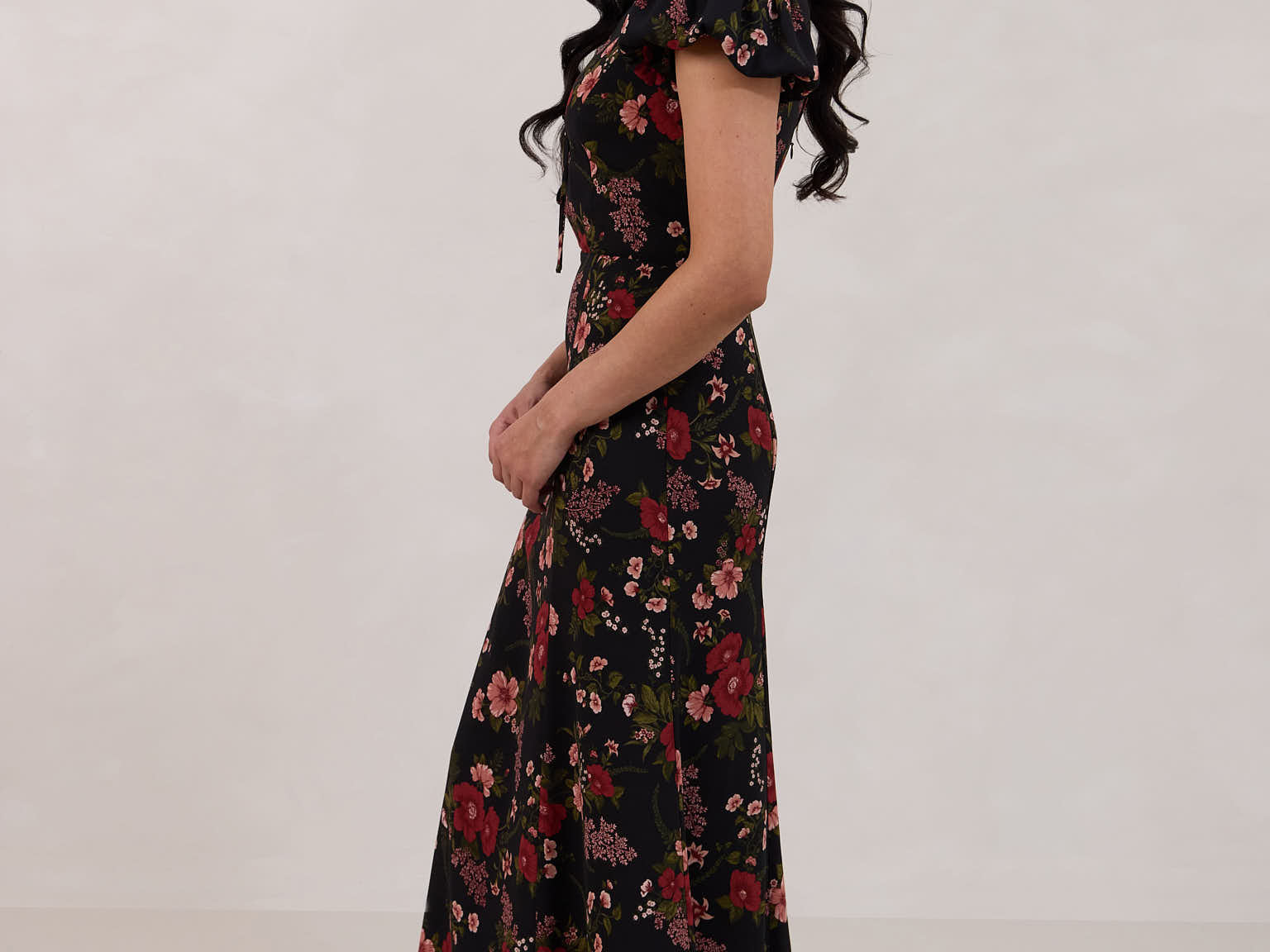 A woman is wearing the Beatrice Dress in Zinnia. This is a black dress with a beautiful red and pink floral print. The dress has a slightly puffed sleeve, and a full back with a keyhole detail. The front has a deep-V cut with a delicate tie at the bust and another keyhole detail down to the waistline. This is a bra-friendly dress that is ankle length. 