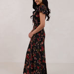 A woman is wearing the Beatrice Dress in Zinnia. This is a black dress with a beautiful red and pink floral print. The dress has a slightly puffed sleeve, and a full back with a keyhole detail. The front has a deep-V cut with a delicate tie at the bust and another keyhole detail down to the waistline. This is a bra-friendly dress that is ankle length. 