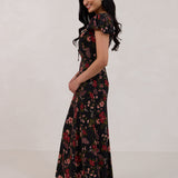 A woman is wearing the Beatrice Dress in Zinnia. This is a black dress with a beautiful red and pink floral print. The dress has a slightly puffed sleeve, and a full back with a keyhole detail. The front has a deep-V cut with a delicate tie at the bust and another keyhole detail down to the waistline. This is a bra-friendly dress that is ankle length. 