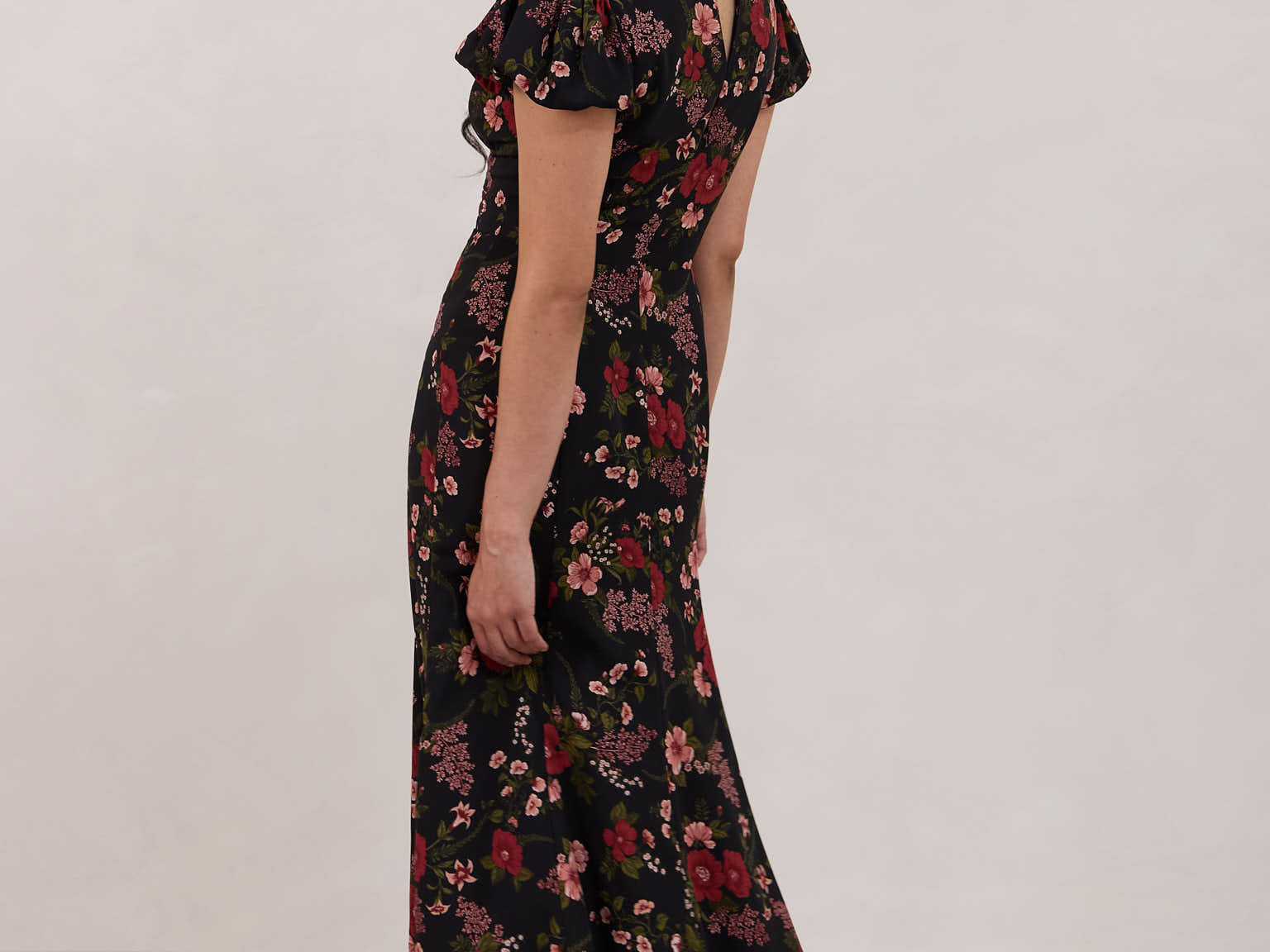 A woman is wearing the Beatrice Dress in Zinnia. This is a black dress with a beautiful red and pink floral print. The dress has a slightly puffed sleeve, and a full back with a keyhole detail. The front has a deep-V cut with a delicate tie at the bust and another keyhole detail down to the waistline. This is a bra-friendly dress that is ankle length. 