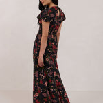 A woman is wearing the Beatrice Dress in Zinnia. This is a black dress with a beautiful red and pink floral print. The dress has a slightly puffed sleeve, and a full back with a keyhole detail. The front has a deep-V cut with a delicate tie at the bust and another keyhole detail down to the waistline. This is a bra-friendly dress that is ankle length. 