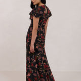A woman is wearing the Beatrice Dress in Zinnia. This is a black dress with a beautiful red and pink floral print. The dress has a slightly puffed sleeve, and a full back with a keyhole detail. The front has a deep-V cut with a delicate tie at the bust and another keyhole detail down to the waistline. This is a bra-friendly dress that is ankle length. 