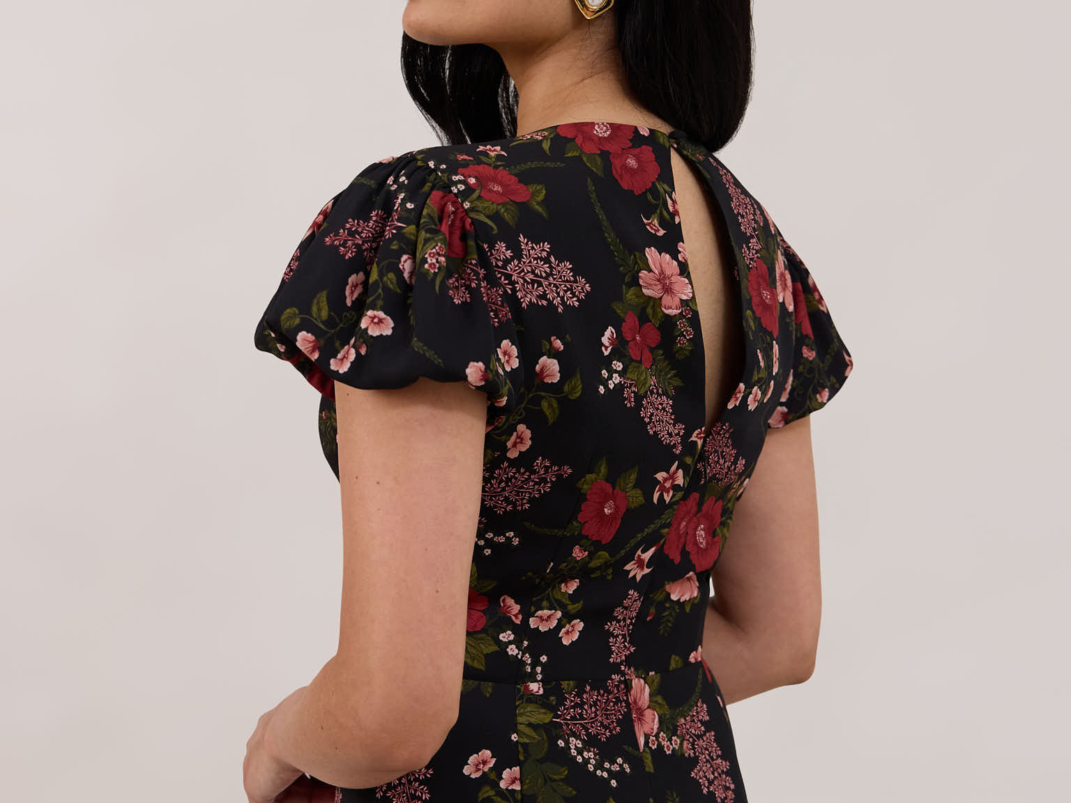 A woman is wearing the Beatrice Dress in Zinnia. This is a black dress with a beautiful red and pink floral print. The dress has a slightly puffed sleeve, and a full back with a keyhole detail. The front has a deep-V cut with a delicate tie at the bust and another keyhole detail down to the waistline. This is a bra-friendly dress that is ankle length. 