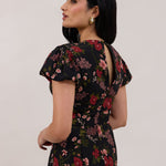 A woman is wearing the Beatrice Dress in Zinnia. This is a black dress with a beautiful red and pink floral print. The dress has a slightly puffed sleeve, and a full back with a keyhole detail. The front has a deep-V cut with a delicate tie at the bust and another keyhole detail down to the waistline. This is a bra-friendly dress that is ankle length. 
