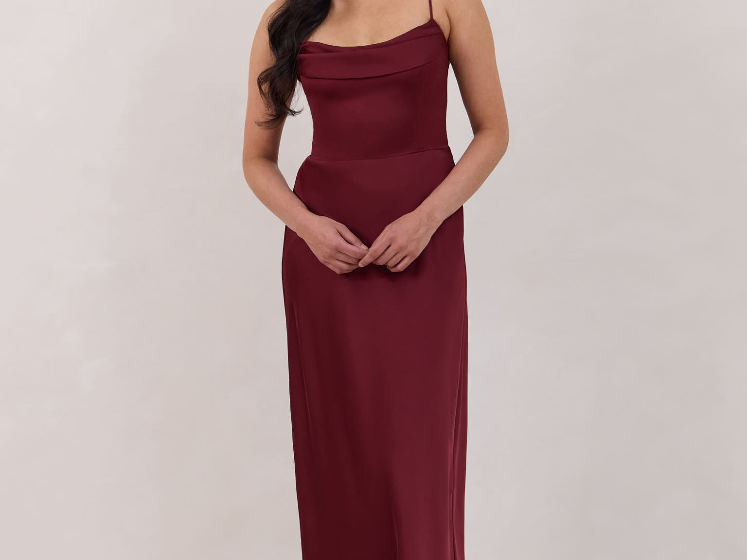 A woman is standing against a white backdrop wearing a burgundy, tea length satin bridesmaid dress. The dress has a boned bodice with a slight gathering at the bust, and a bias cut skirt that falls nicely over the hips. This is the Rosedale dress in merlot.