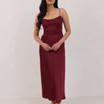 A woman is standing against a white backdrop wearing a burgundy, tea length satin bridesmaid dress. The dress has a boned bodice with a slight gathering at the bust, and a bias cut skirt that falls nicely over the hips. This is the Rosedale dress in merlot.