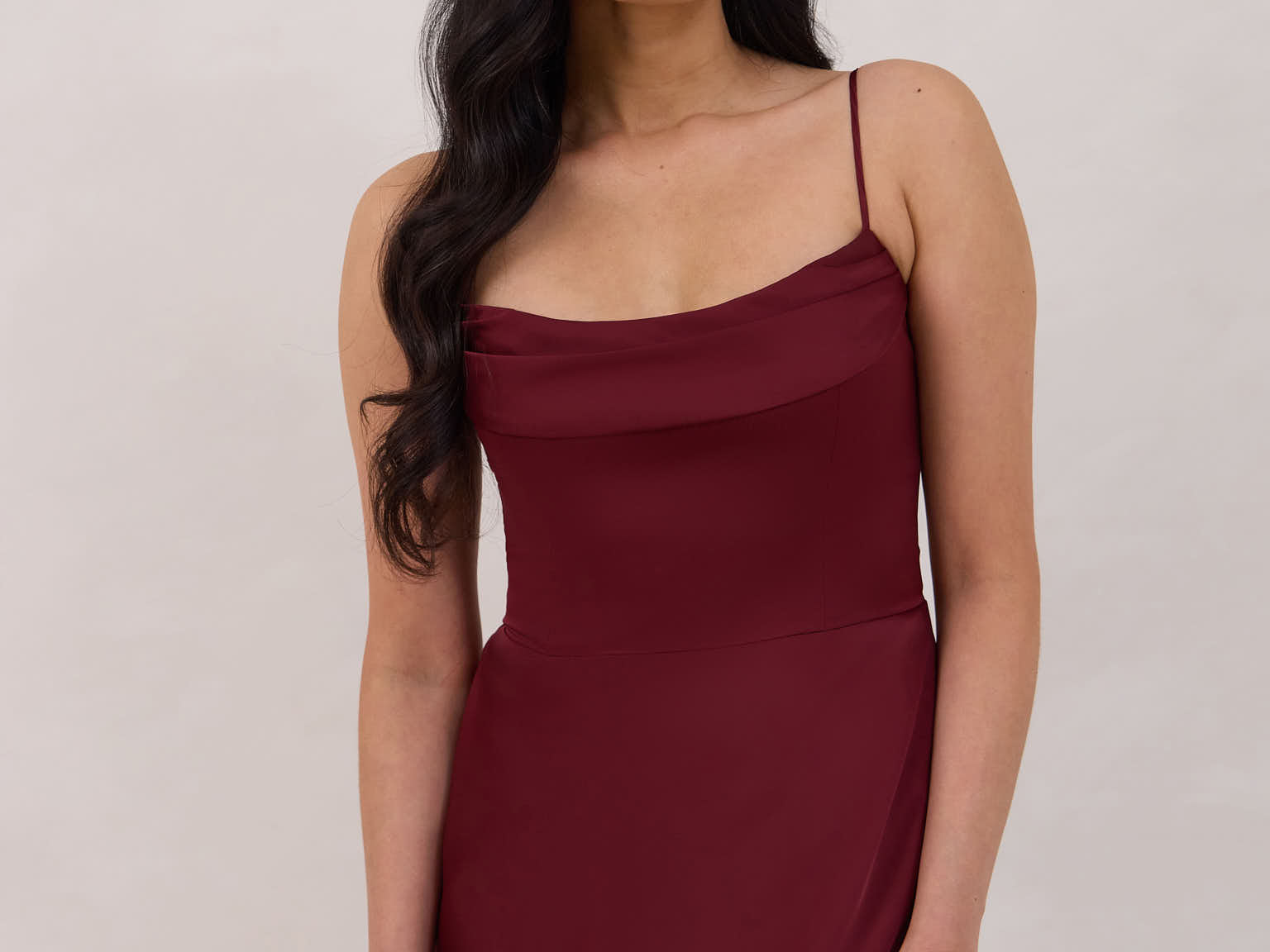 A woman is standing against a white backdrop wearing a burgundy, tea length satin bridesmaid dress. The dress has a boned bodice with a slight gathering at the bust, and a bias cut skirt that falls nicely over the hips. This is the Rosedale dress in merlot.