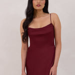 A woman is standing against a white backdrop wearing a burgundy, tea length satin bridesmaid dress. The dress has a boned bodice with a slight gathering at the bust, and a bias cut skirt that falls nicely over the hips. This is the Rosedale dress in merlot.
