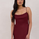 A woman is standing against a white backdrop wearing a burgundy, tea length satin bridesmaid dress. The dress has a boned bodice with a slight gathering at the bust, and a bias cut skirt that falls nicely over the hips. This is the Rosedale dress in merlot.