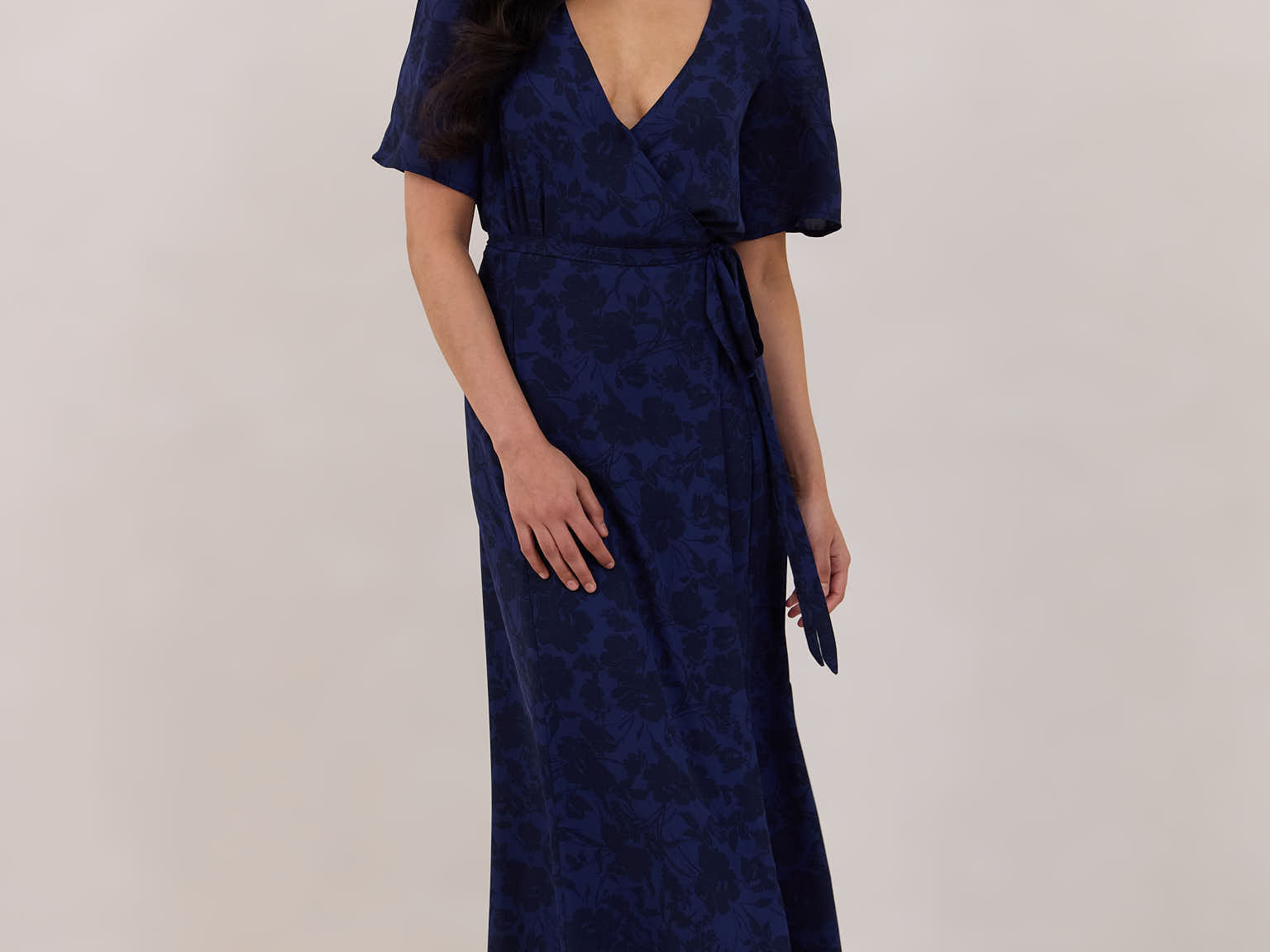 A woman is standing against a white backdrop wearing a navy blue wrap dress with a subtle black floral print. The dress has a deep V neckline and loose sleeves. This is they Sherwood Dress in Twyla.