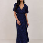 A woman is standing against a white backdrop wearing a navy blue wrap dress with a subtle black floral print. The dress has a deep V neckline and loose sleeves. This is they Sherwood Dress in Twyla.