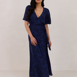 A woman is standing against a white backdrop wearing a navy blue wrap dress with a subtle black floral print. The dress has a deep V neckline and loose sleeves. This is they Sherwood Dress in Twyla.