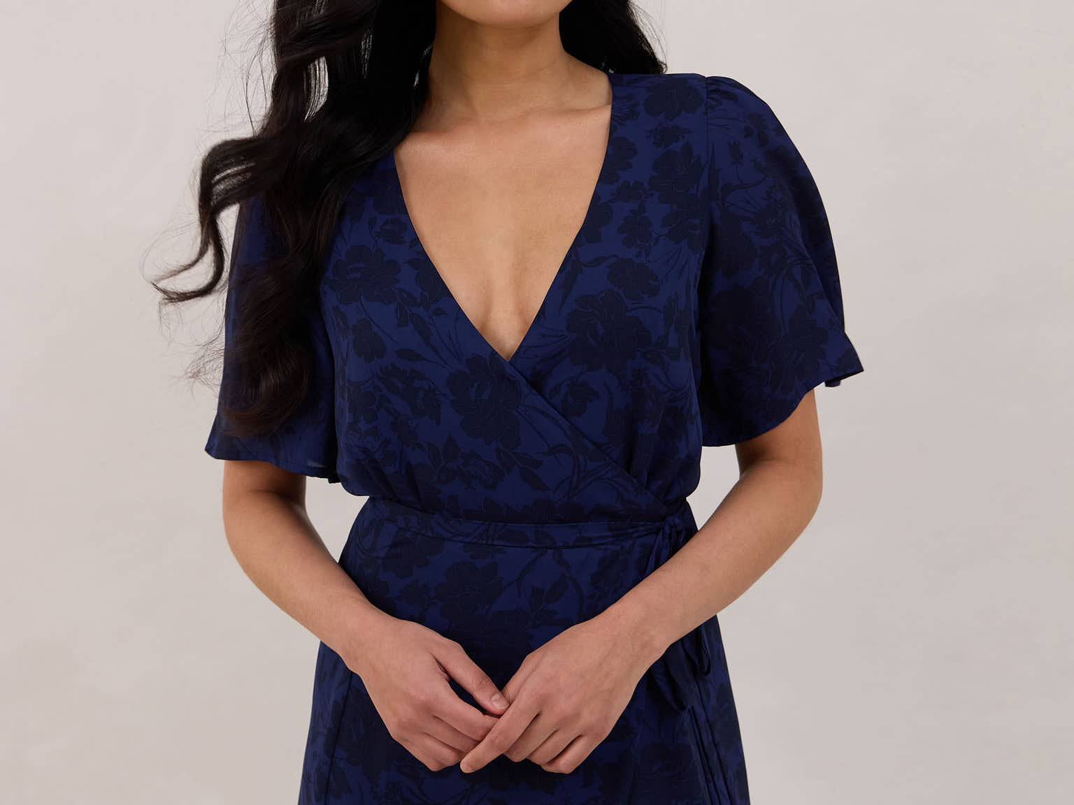 A woman is standing against a white backdrop wearing a navy blue wrap dress with a subtle black floral print. The dress has a deep V neckline and loose sleeves. This is they Sherwood Dress in Twyla.