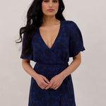 A woman is standing against a white backdrop wearing a navy blue wrap dress with a subtle black floral print. The dress has a deep V neckline and loose sleeves. This is they Sherwood Dress in Twyla.
