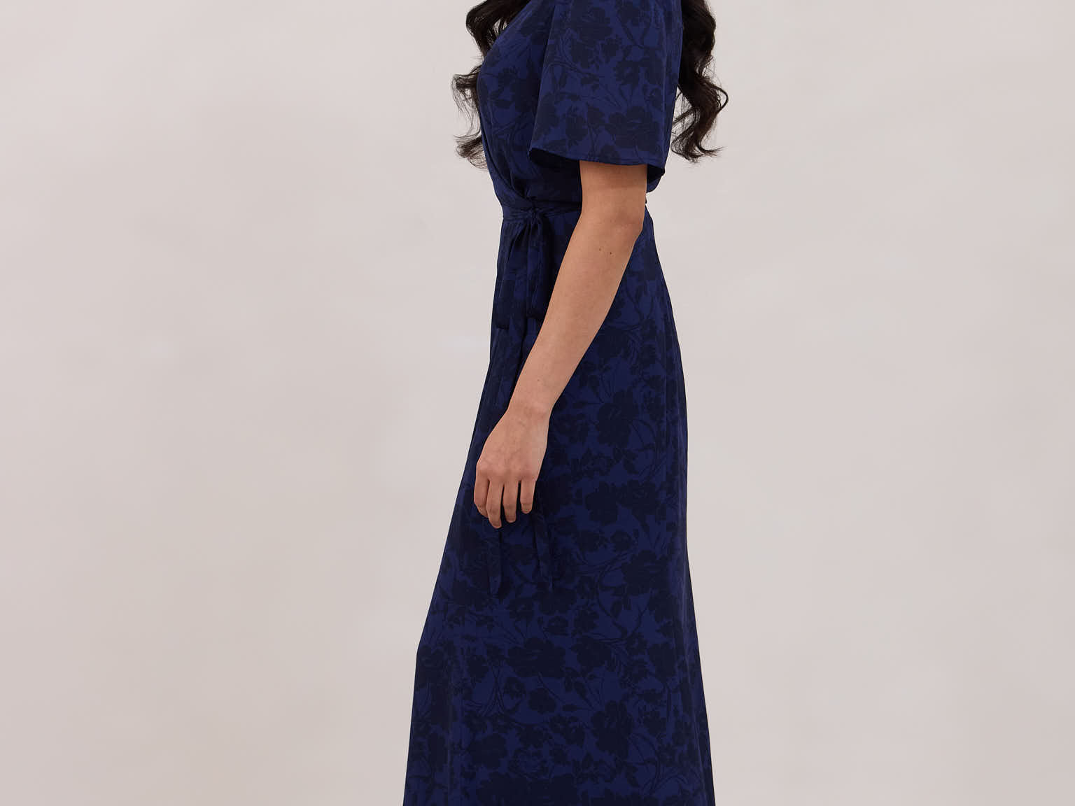 A woman is standing against a white backdrop facing the left wearing a navy blue wrap dress with a subtle black floral print. The dress has a deep V neckline and loose sleeves. This is they Sherwood Dress in Twyla.