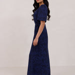 A woman is standing against a white backdrop facing the left wearing a navy blue wrap dress with a subtle black floral print. The dress has a deep V neckline and loose sleeves. This is they Sherwood Dress in Twyla.