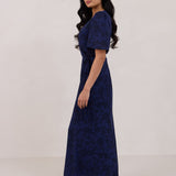A woman is standing against a white backdrop facing the left wearing a navy blue wrap dress with a subtle black floral print. The dress has a deep V neckline and loose sleeves. This is they Sherwood Dress in Twyla.