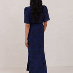 A woman is standing against a white backdrop facing the back wearing a navy blue wrap dress with a subtle black floral print. The dress has a deep V neckline and loose sleeves. This is they Sherwood Dress in Twyla.