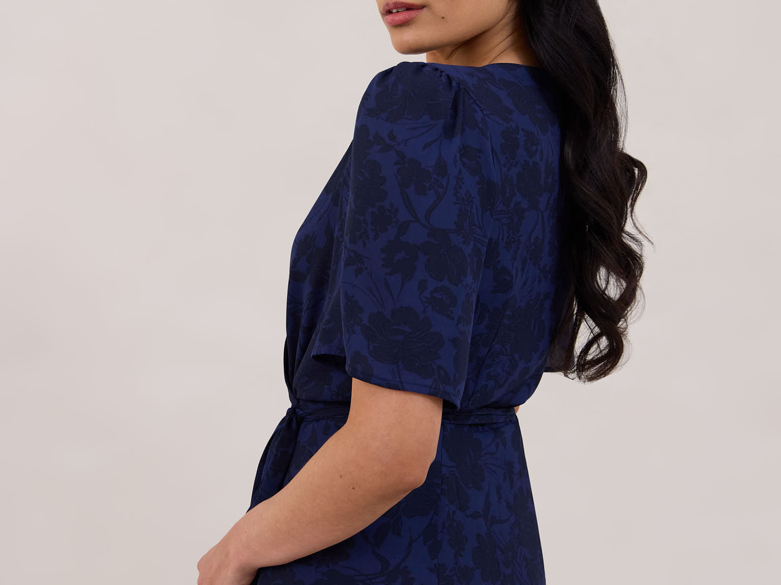A woman is standing against a white backdrop facing the back and looking over her shoulder wearing a navy blue wrap dress with a subtle black floral print. The dress has a deep V neckline and loose sleeves. This is they Sherwood Dress in Twyla.