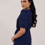 A woman is standing against a white backdrop facing the back and looking over her shoulder wearing a navy blue wrap dress with a subtle black floral print. The dress has a deep V neckline and loose sleeves. This is they Sherwood Dress in Twyla.