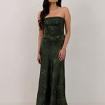 A woman standing against a white backdrop wearing a strapless tea length dress. The dress is a dark green with a subtle green floral print. The dress is fitted through the bodice, and has a more flowy skirt. This is the Sterling dress in Fern.
