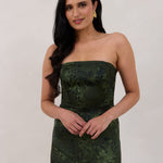 A woman standing against a white backdrop wearing a strapless tea length dress. The dress is a dark green with a subtle green floral print. The dress is fitted through the bodice, and has a more flowy skirt. This is the Sterling dress in Fern.