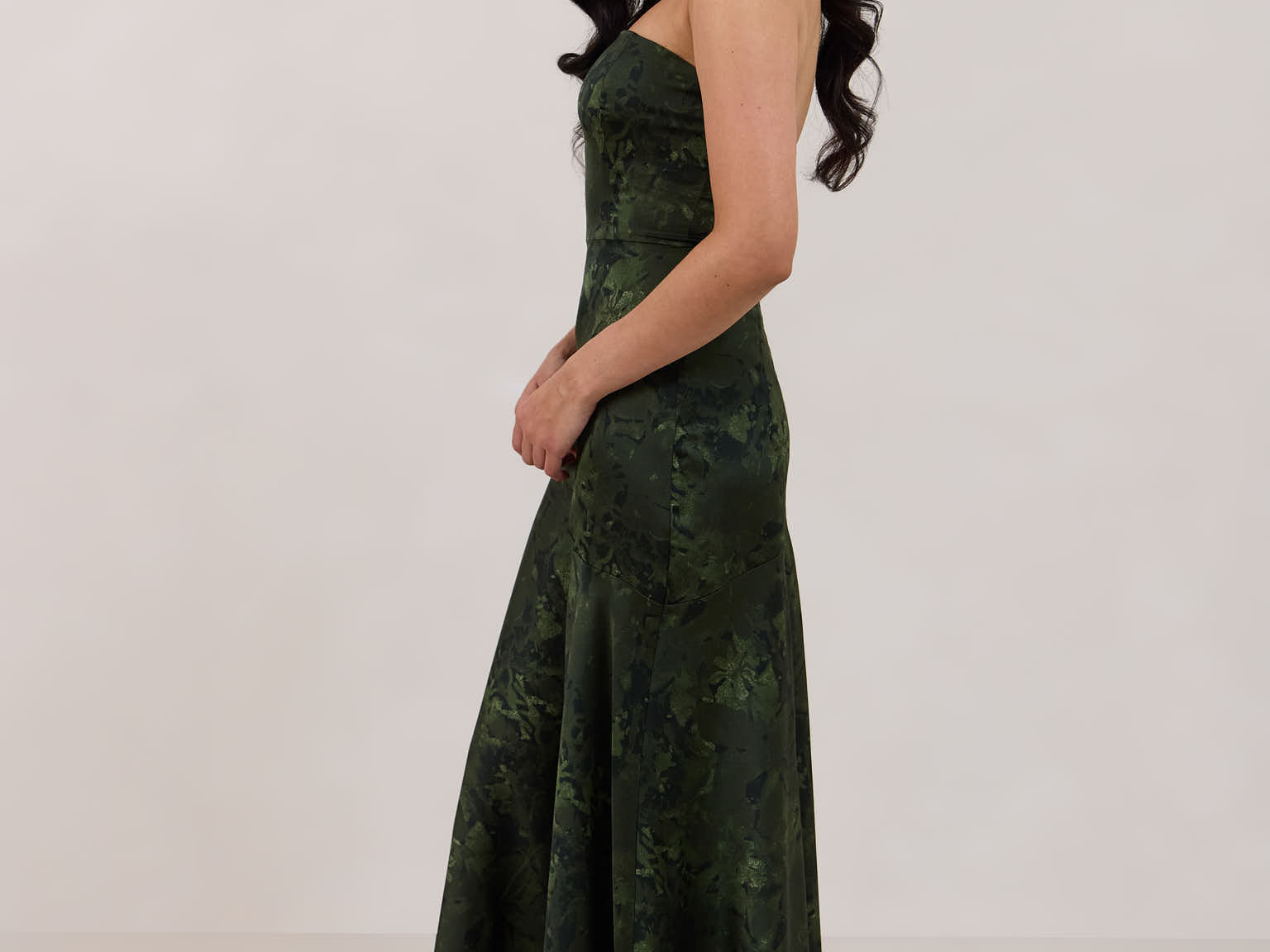 A woman standing facing the left against a white backdrop wearing a strapless tea length dress. The dress is a dark green with a subtle green floral print. The dress is fitted through the bodice, and has a more flowy skirt. This is the Sterling dress in Fern.