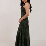 A woman standing facing the left against a white backdrop wearing a strapless tea length dress. The dress is a dark green with a subtle green floral print. The dress is fitted through the bodice, and has a more flowy skirt. This is the Sterling dress in Fern.
