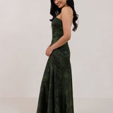 A woman standing facing the left against a white backdrop wearing a strapless tea length dress. The dress is a dark green with a subtle green floral print. The dress is fitted through the bodice, and has a more flowy skirt. This is the Sterling dress in Fern.