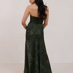 A woman standing facing the back against a white backdrop wearing a strapless tea length dress. The dress is a dark green with a subtle green floral print. The dress is fitted through the bodice, and has a more flowy skirt. This is the Sterling dress in Fern.