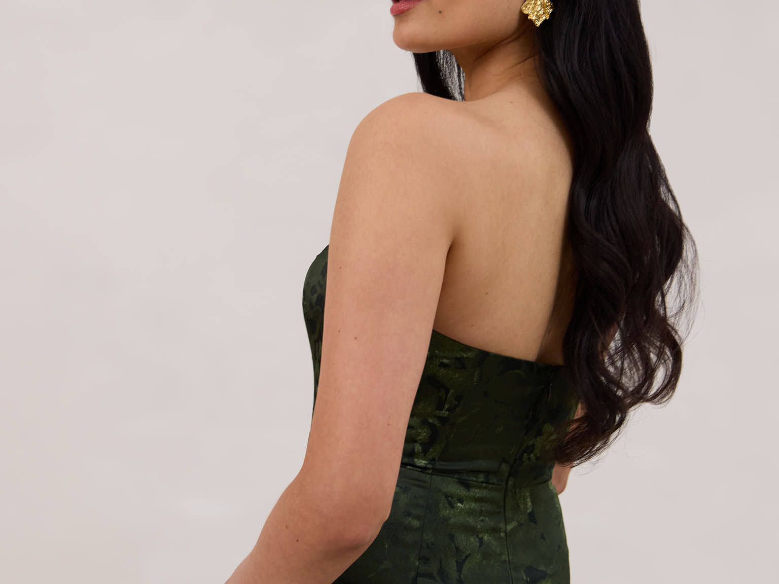 A woman standing facing the back against a white backdrop wearing a strapless tea length dress. The dress is a dark green with a subtle green floral print. The dress is fitted through the bodice, and has a more flowy skirt. This is the Sterling dress in Fern.