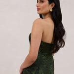A woman standing facing the back against a white backdrop wearing a strapless tea length dress. The dress is a dark green with a subtle green floral print. The dress is fitted through the bodice, and has a more flowy skirt. This is the Sterling dress in Fern.