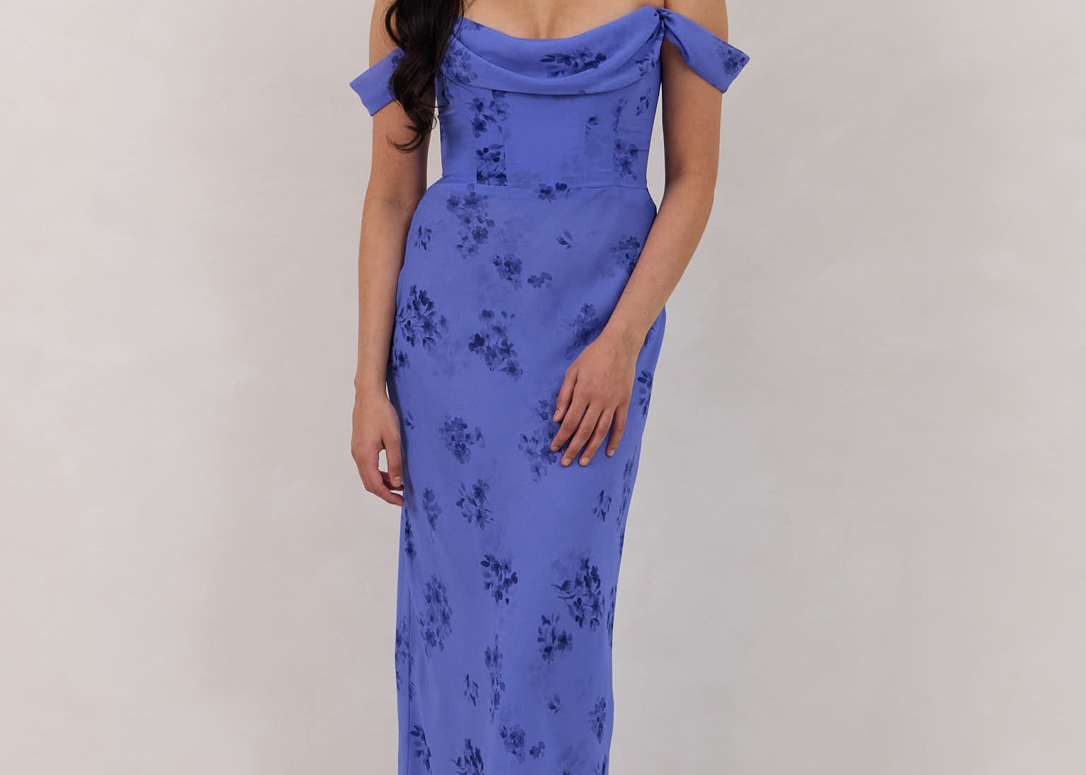 A woman standing against a white background wearing a periwinkle blue floral print dress. The dress has a scooped neckline with an off-the-shoulder strap, is fitted through the bodice and falls to ankle length. The fabric is chiffon. This is the Sutton Dress in Livia. 