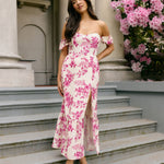 A woman standing on outdoor patio steps. She's wearing a cream coloured dress with a pink floral print. The dress has a strapless sweetheart neckline with an off-the-shoulder sleeve. The skirt has a thigh high slit and a ruffle detail along the hem. This is the Bentley Dress in Mulberry.