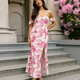 A woman standing on outdoor patio steps. She's wearing a cream coloured dress with a pink floral print. The dress has a strapless sweetheart neckline with an off-the-shoulder sleeve. The skirt has a thigh high slit and a ruffle detail along the hem. This is the Bentley Dress in Mulberry.